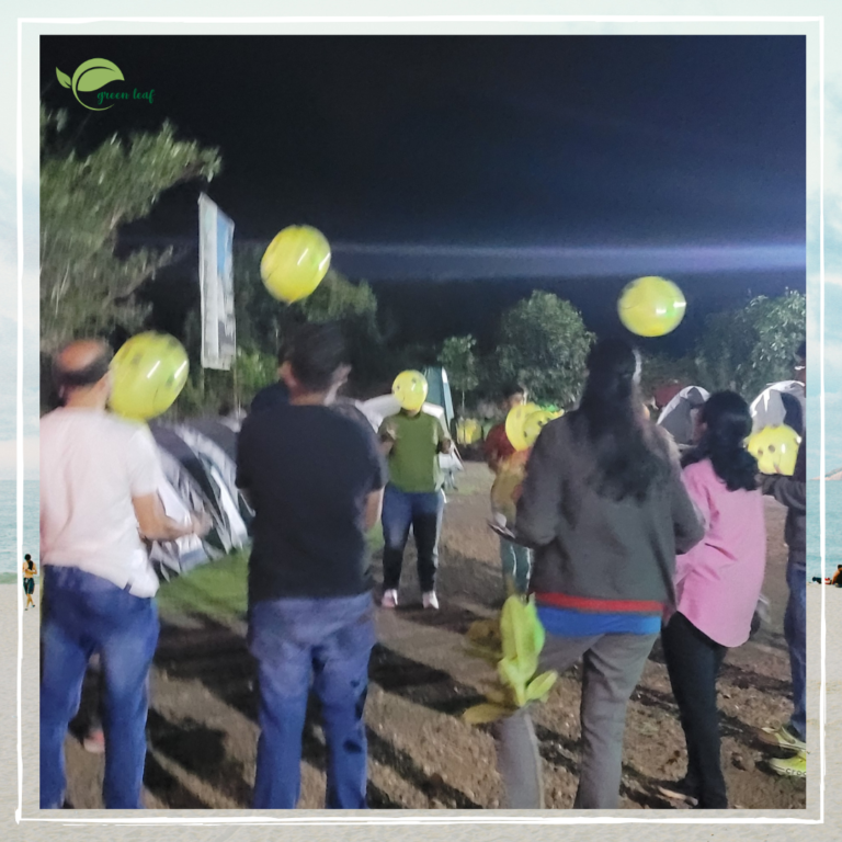 "Group of people playing with yellow smiley face balloons at night during a camping trip near Pune, with tents and trees in the background."